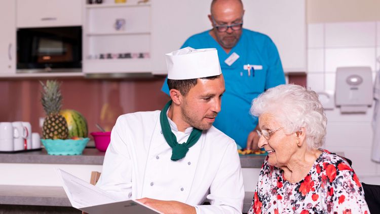 Koch mit Kochmütze serviert Seniorin Essen